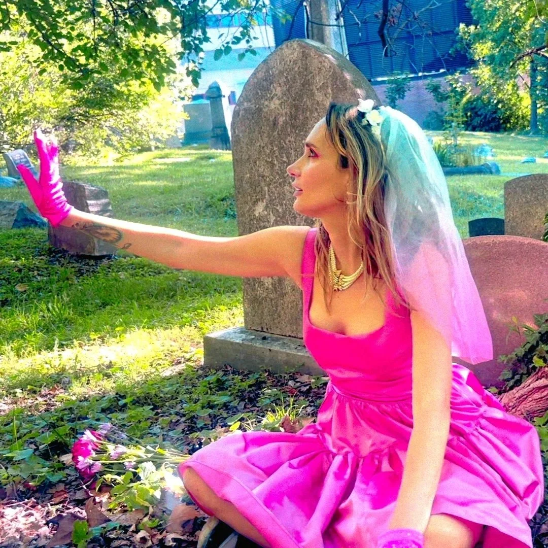 A woman dressed as a bride with a pink dress, veil, and yellow necklace sitting on the grass in a cemetery, reaching towards a flower on the ground with a pink glove.