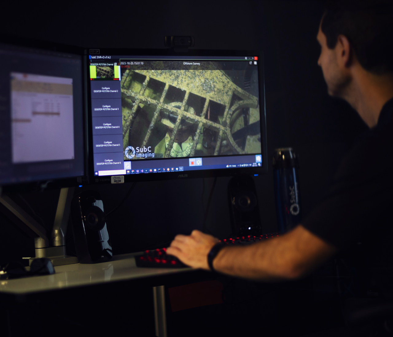 Person sitting at a desk with two computer monitors, one showing subsea imaging footage of an underwater structure with the SubC Imaging logo.