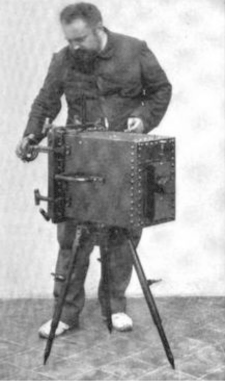 Man standing behind a square-shaped camera on a tripod.