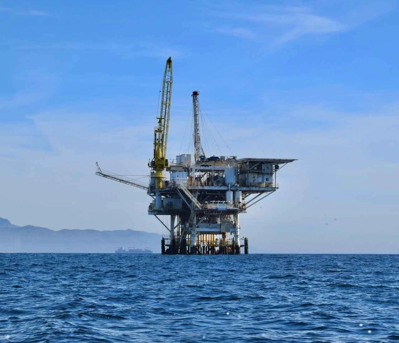 Offshore oil rig structure with cranes standing in the ocean under a clear blue sky, with distant mountains faintly visible on the horizon