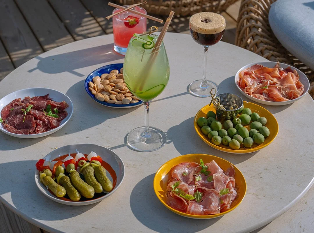 Ett runt bord med olika snacks och drycker, inklusive skivor av kött, charkuterier, oliver, små gurkor, jordgubbar, drinkar och ett glas rödvin.