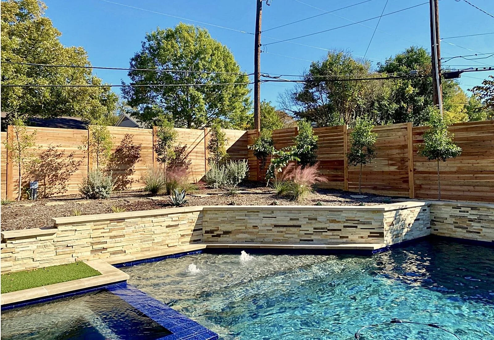 Backyard with a blue swimming pool, wooden privacy fence, trees, and a vegetable garden bed with plants under a sunny sky.