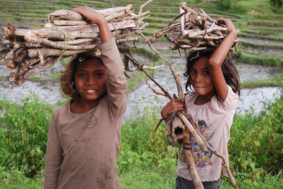 two timorese children smiling cheekily with bundles of sticks on their head in front of a rice field