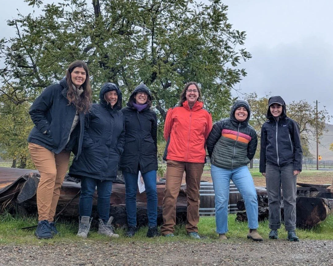 Six smiling people pose for a photo standing in front of a large log wearing raincoats.