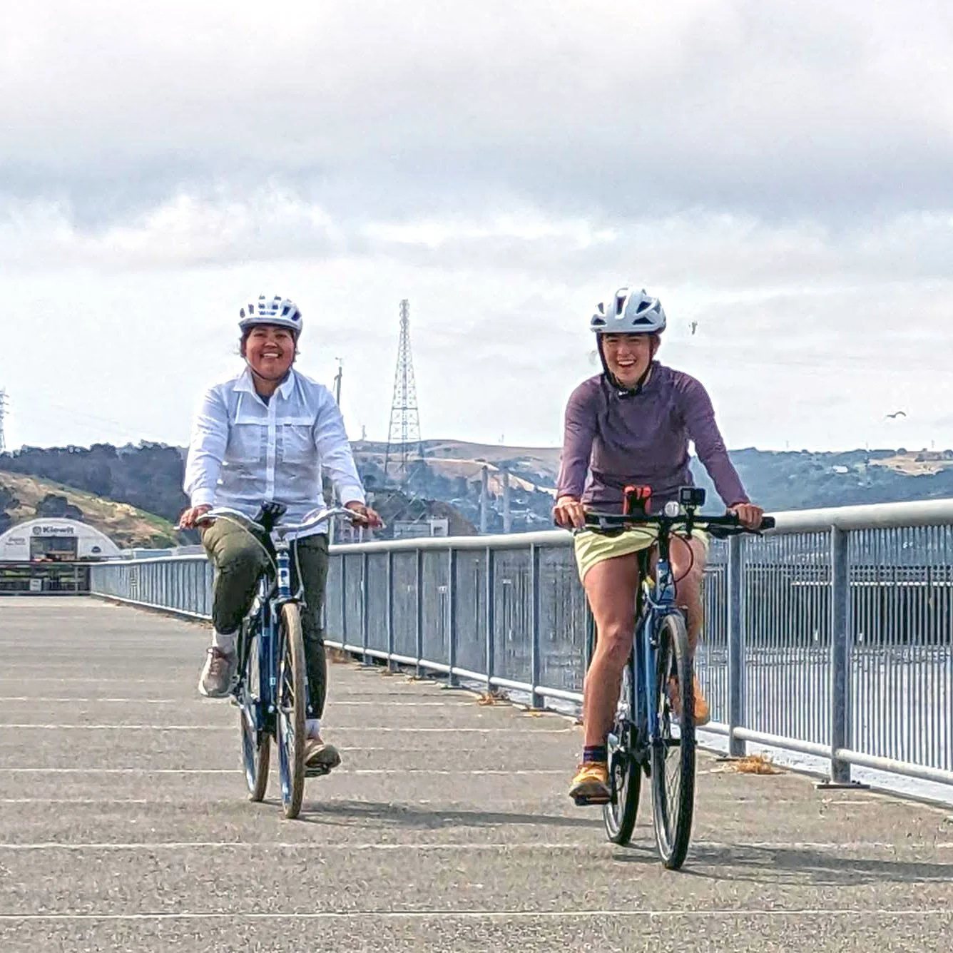 Two women biking along a bike path with mountains in the background and the ocean to the right.