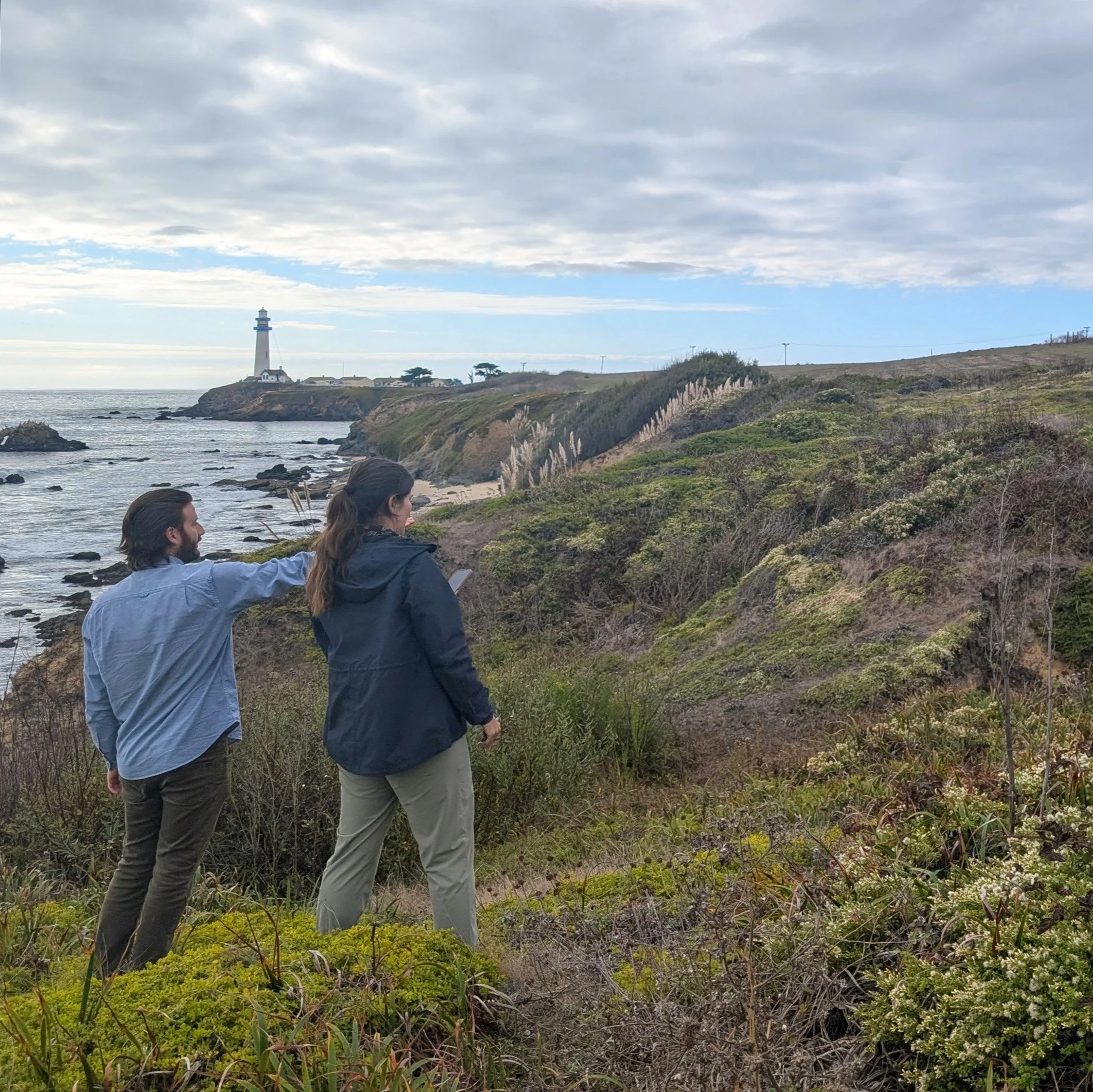 Two people standing and pointing on a coastal bluff with a lighthouse in the background.