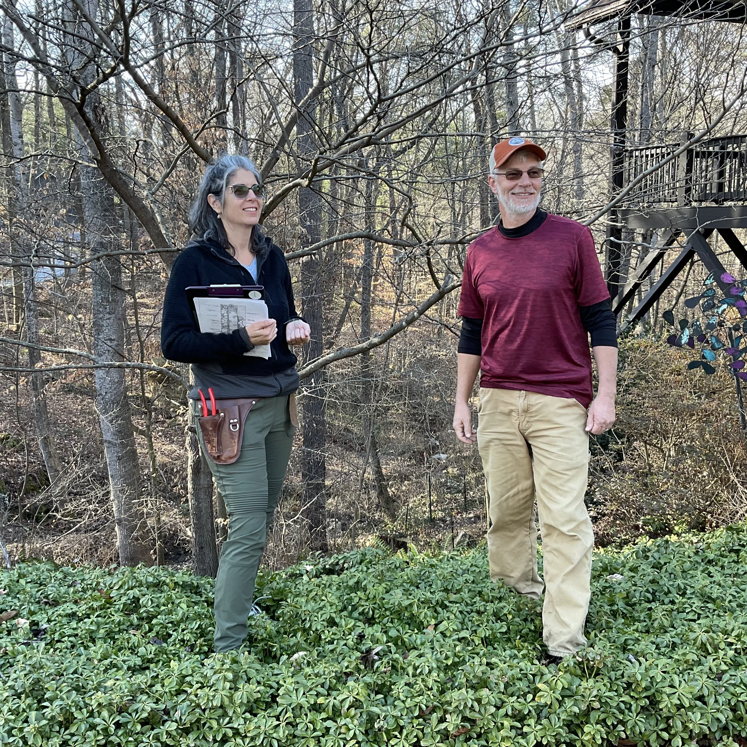 A woman and a man standing outdoors in a wooded area during daytime, smiling and talking, with trees and a wooden staircase in the background.
