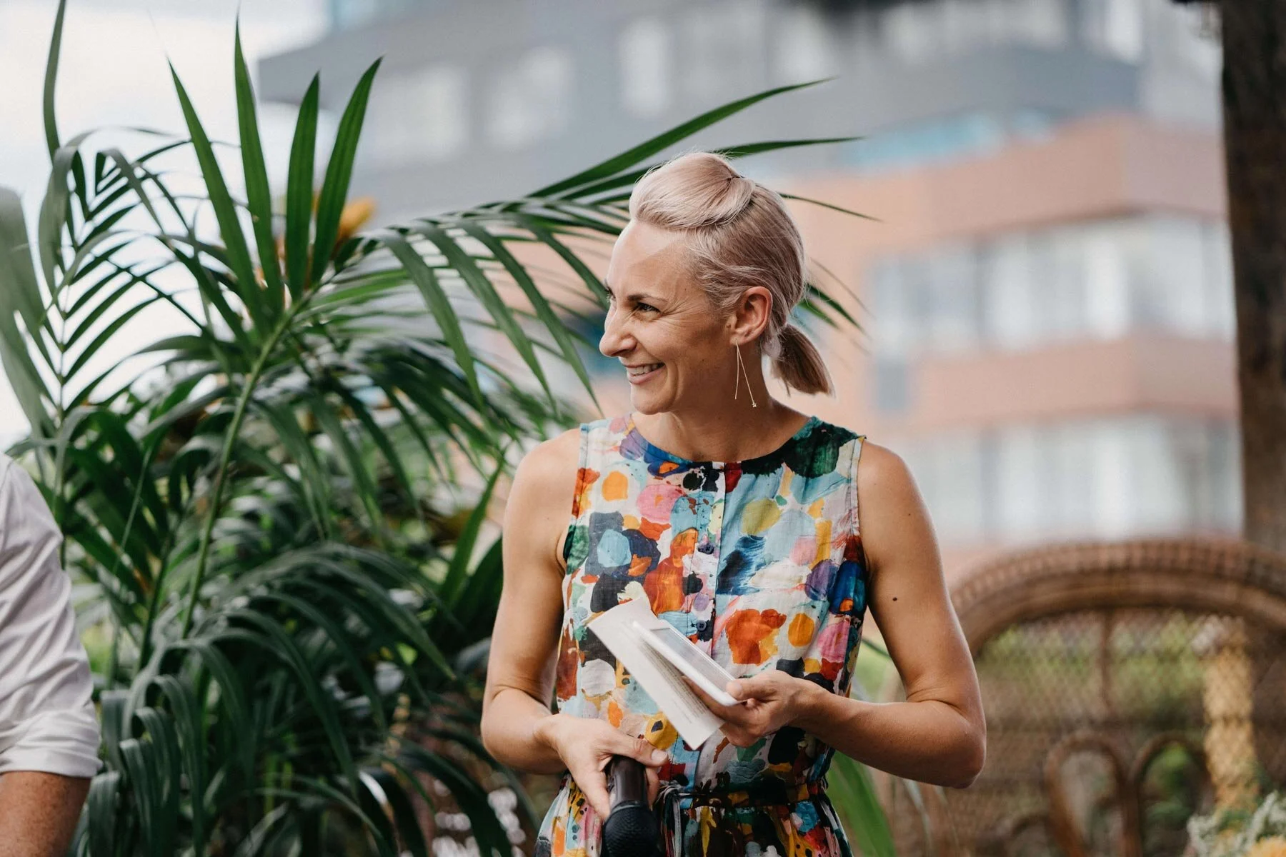 Jacqua Celebrant in multicoloured dress holding her mic and kindle, standing in front of plants ready to conduct a ceremony
