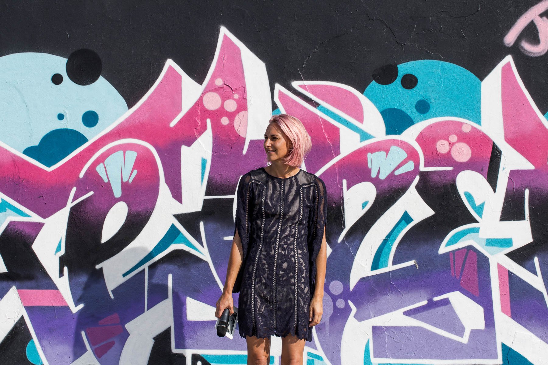 Jacqua Celebrant in navy blue dress holding her mic and kindle, standing in front of a grafitti artwork, Bondi Beach