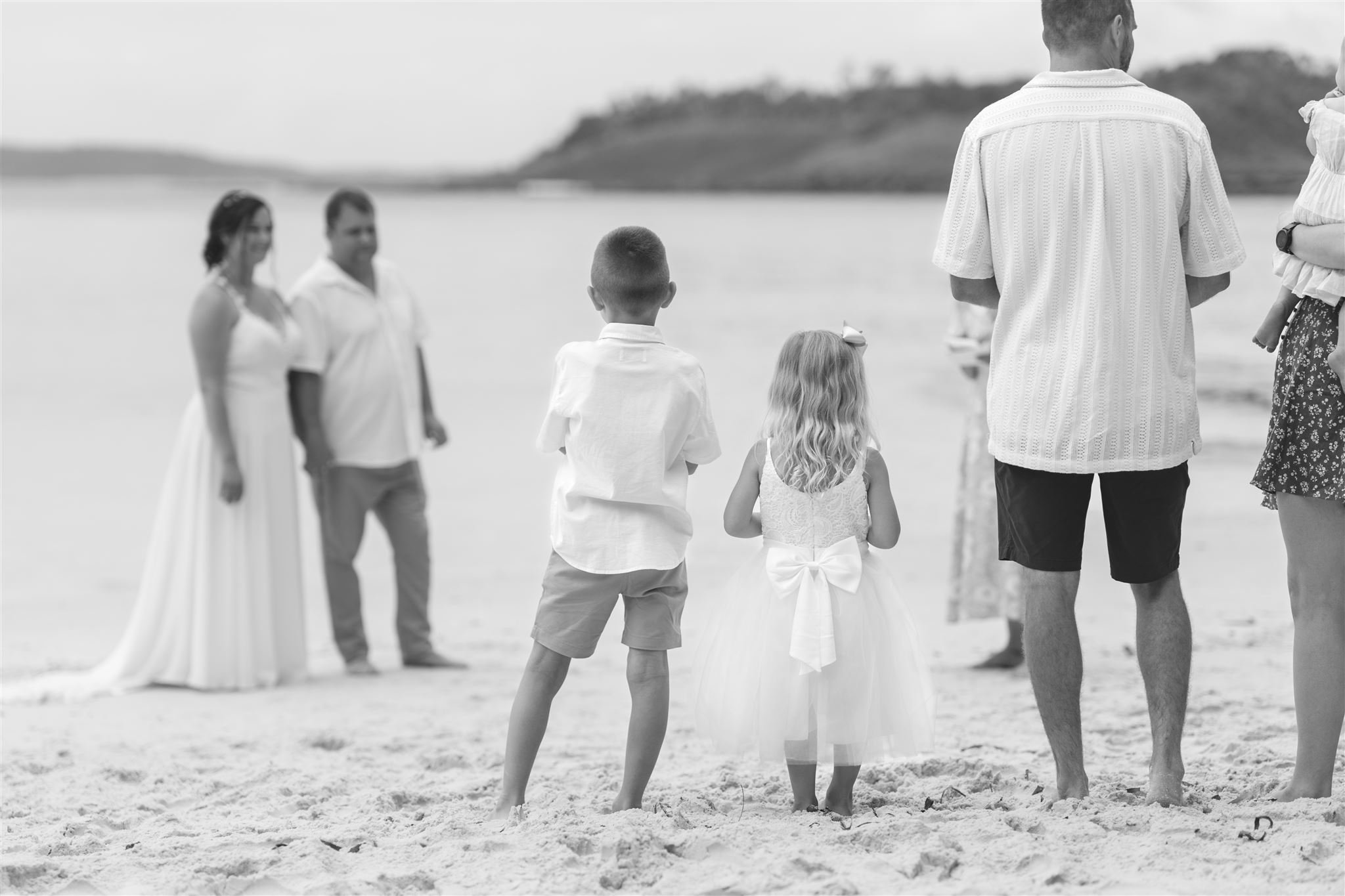 Couple eloping on the shoreline of Murrays Beach, Jervis Bay. Their two children and family members looking on as the ceremony begins.