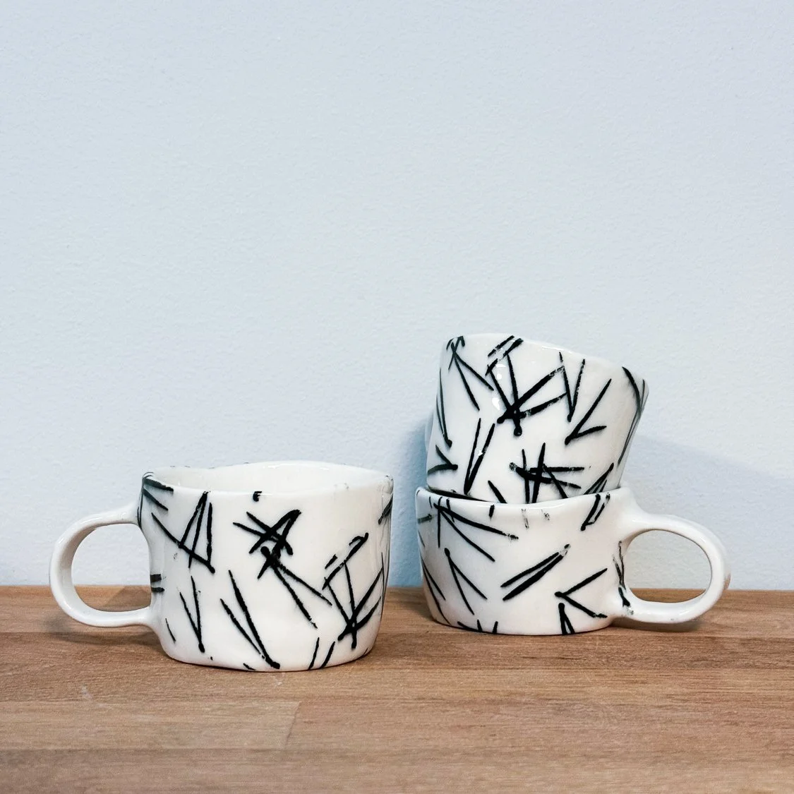 Four white ceramic coffee mugs with black abstract line patterns, stacked and placed on a wooden surface against a plain white wall.