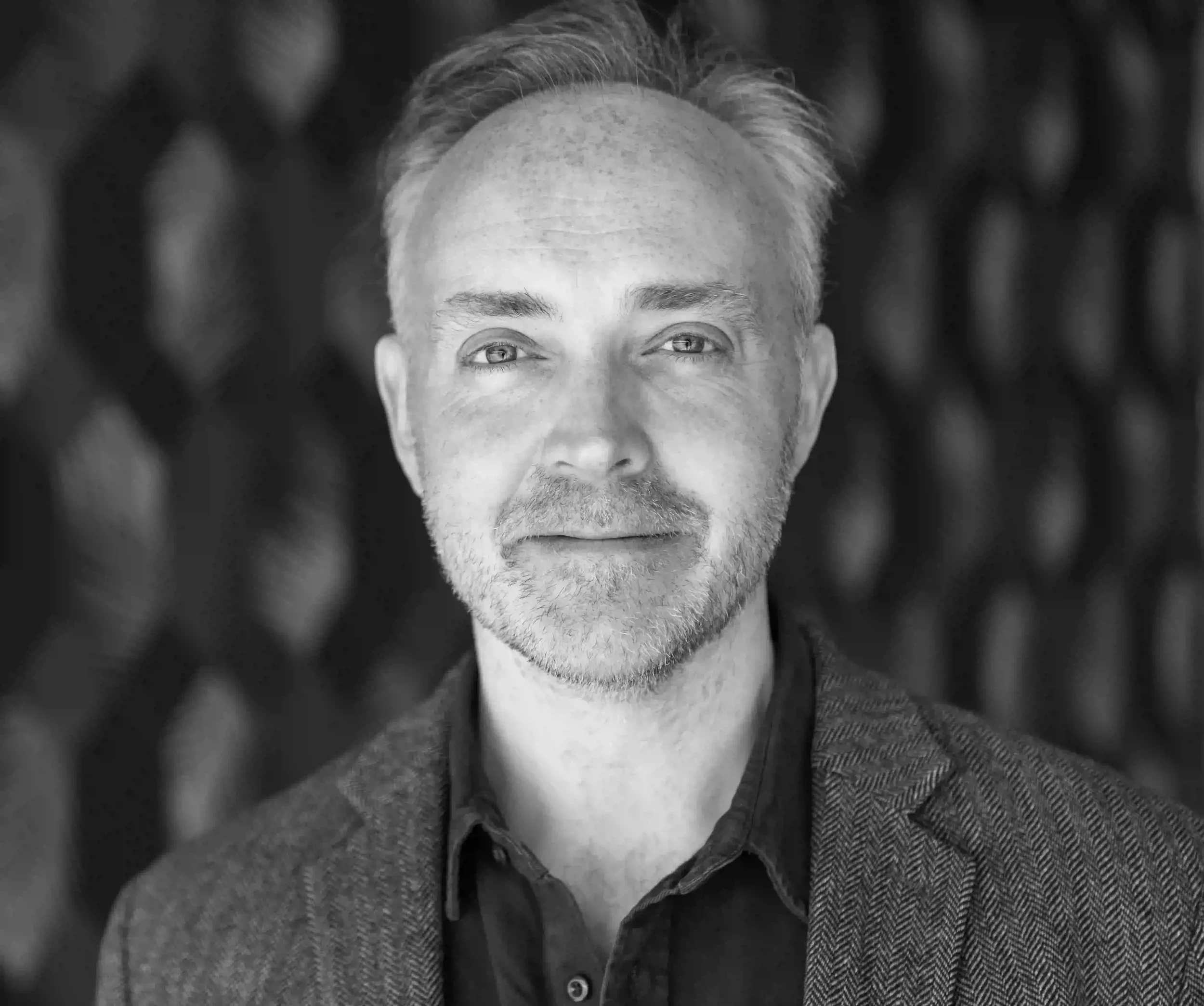 Close-up black and white photograph of a middle-aged man with short gray hair, facial hair, and light-colored eyes, looking directly at the camera with a serious expression, wearing a checkered blazer and a shirt.