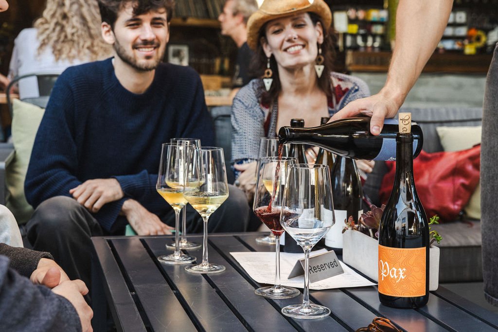 Two smiling people seated at an outdoor table, having wine tasting, with glasses of white and red wine, a bottle of wine labeled 'Pax', and a person pouring red wine.