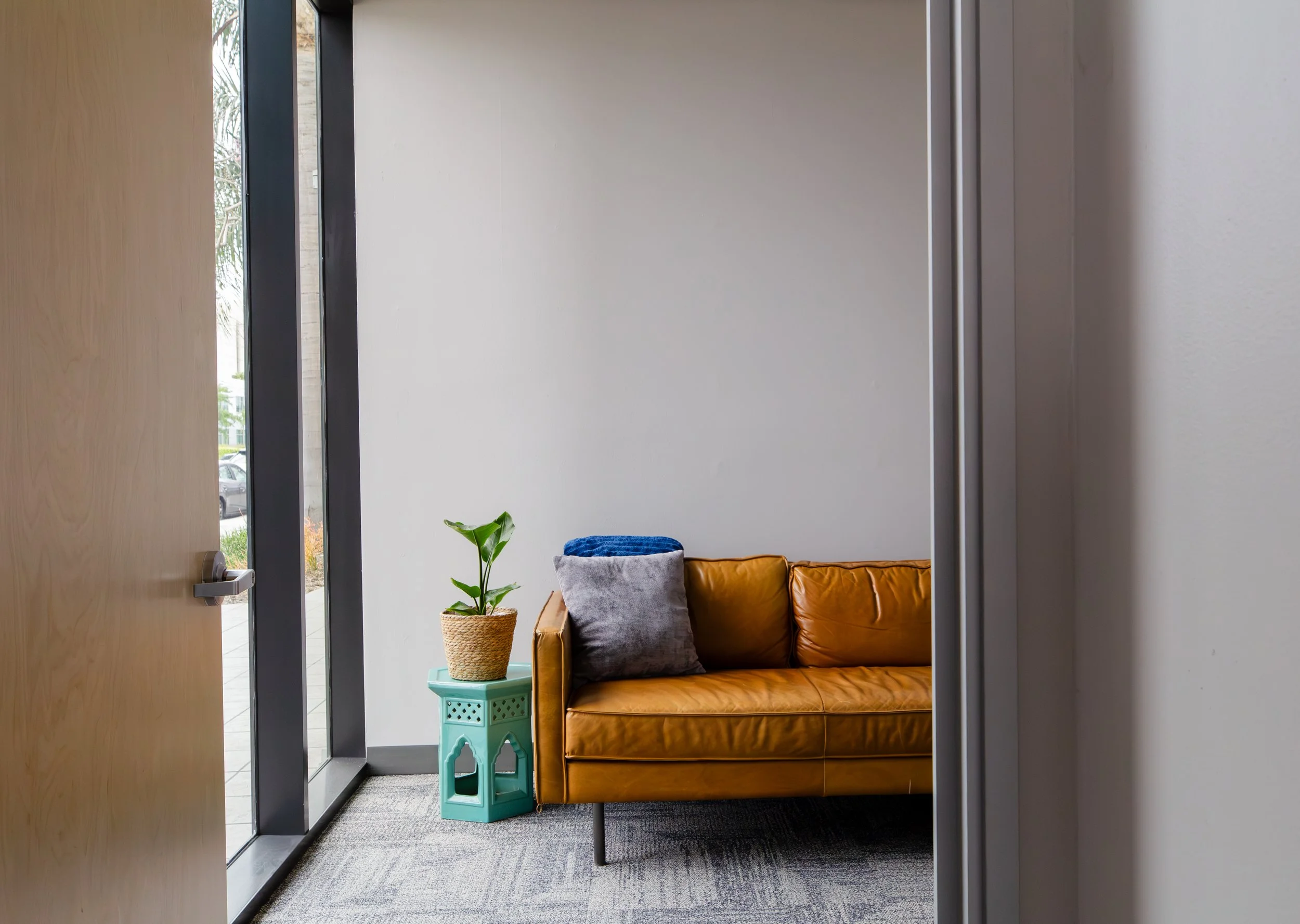 Interior scene of a waiting area with a tan leather sofa topped with gray and blue pillows, a small turquoise side table holding a potted plant, white walls, and large glass windows showing outdoor trees and parked cars. ONA Studios.