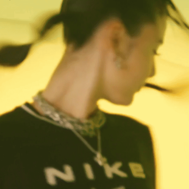 Emei wearing a black Nike shirt and layered necklaces, set against a yellow background.