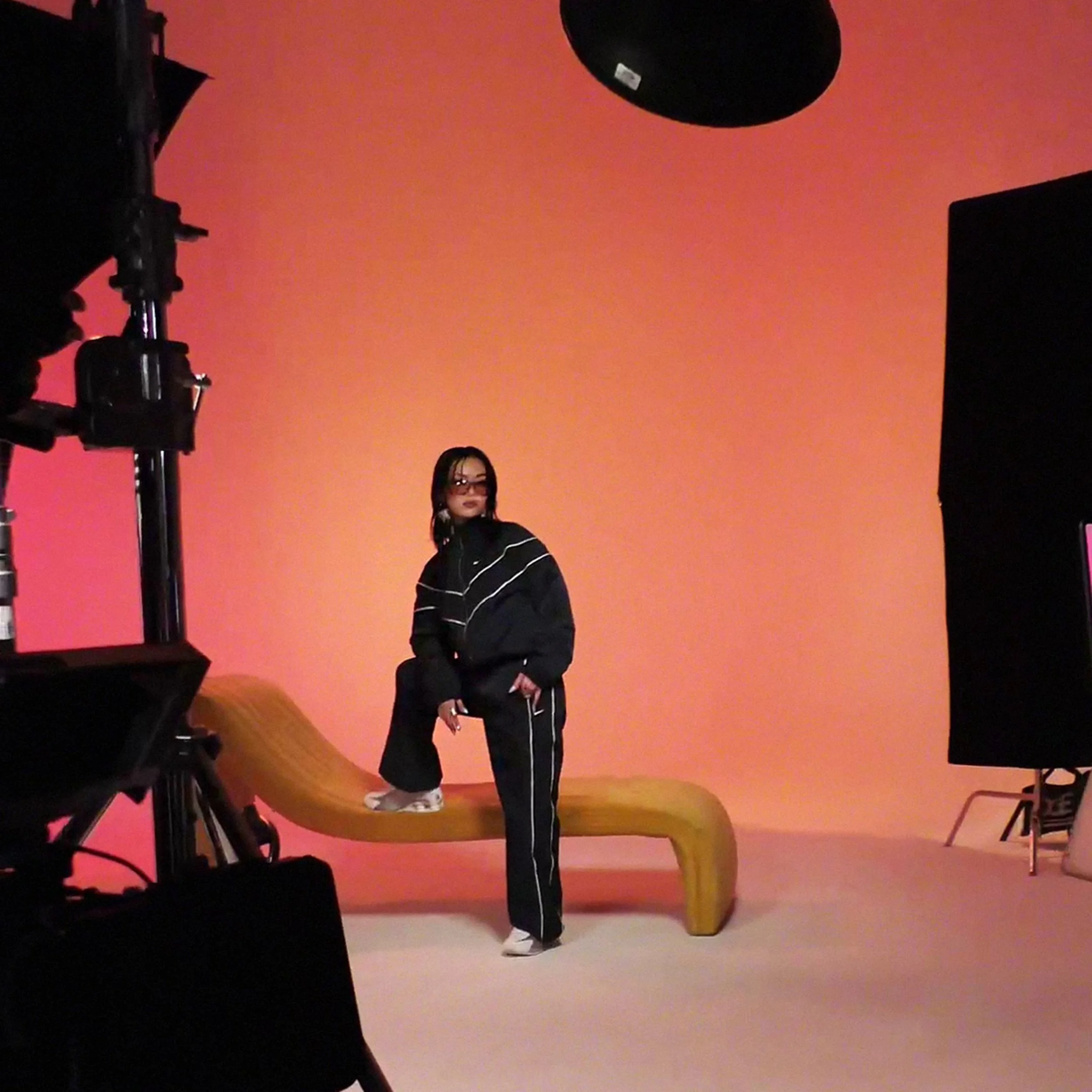 A woman dressed in a black tracksuit with white stripes poses on a wooden bench in front of a peach-colored screen in a photography studio.