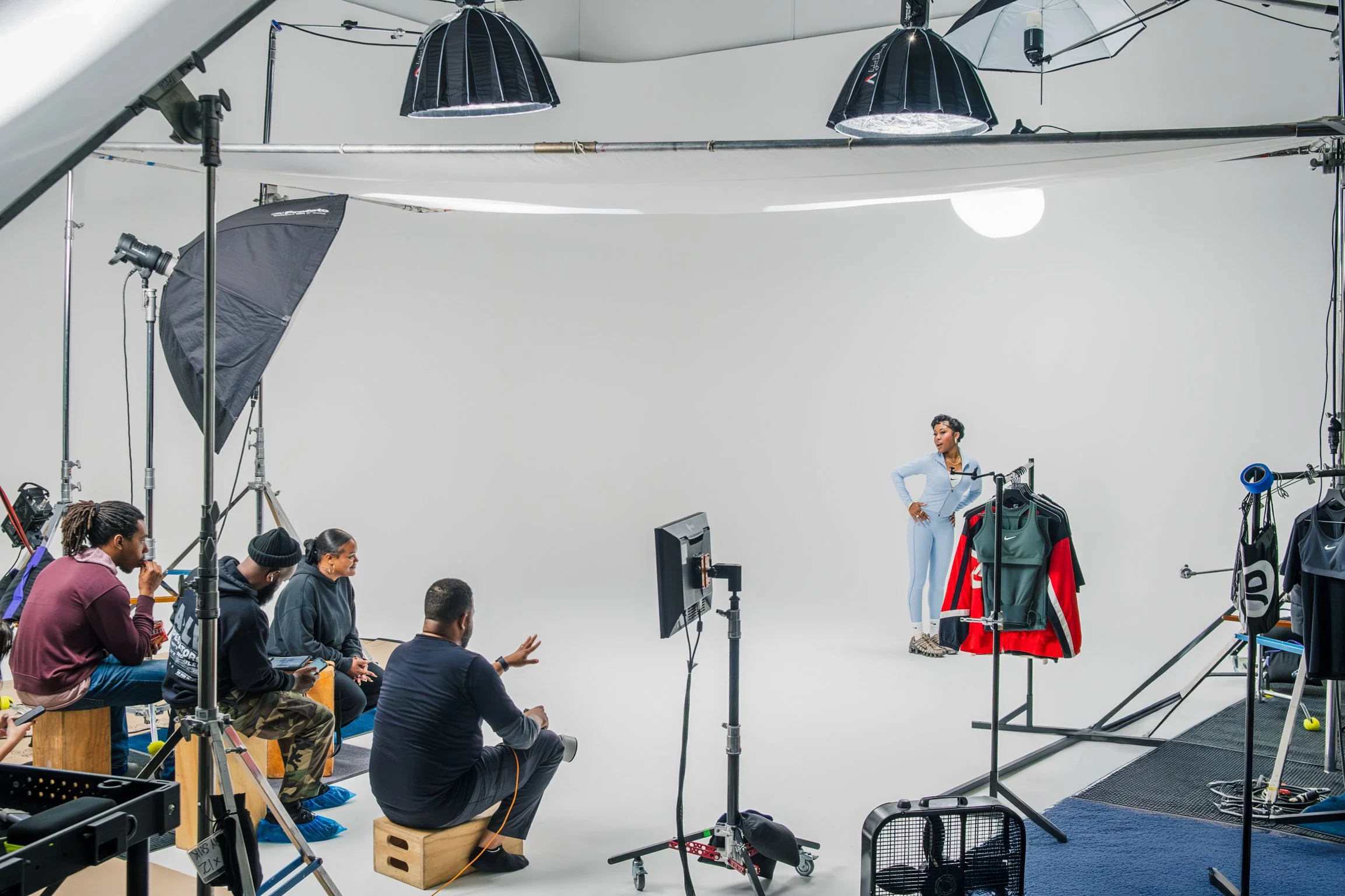 A photoshoot studio with a white background, featuring a woman in light blue athletic wear posing next to clothing racks with colorful sportswear. Several crew members, including a person with a camera, are sitting or standing nearby, coordinating the shoot, with professional lighting equipment overhead.