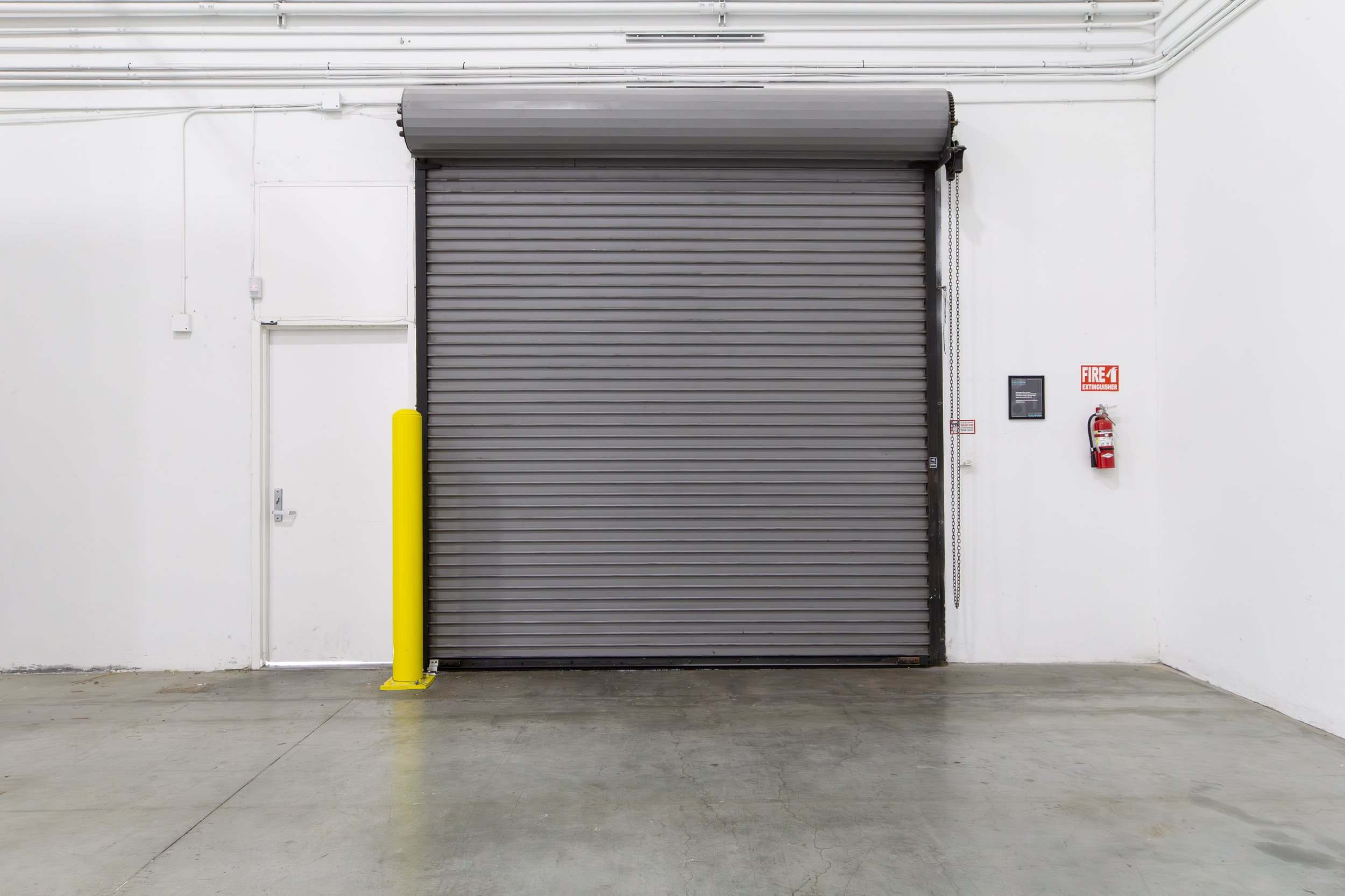 Gray industrial rolling door in a warehouse with a fire extinguisher on the right wall and a small door to the left. ONA Studios.