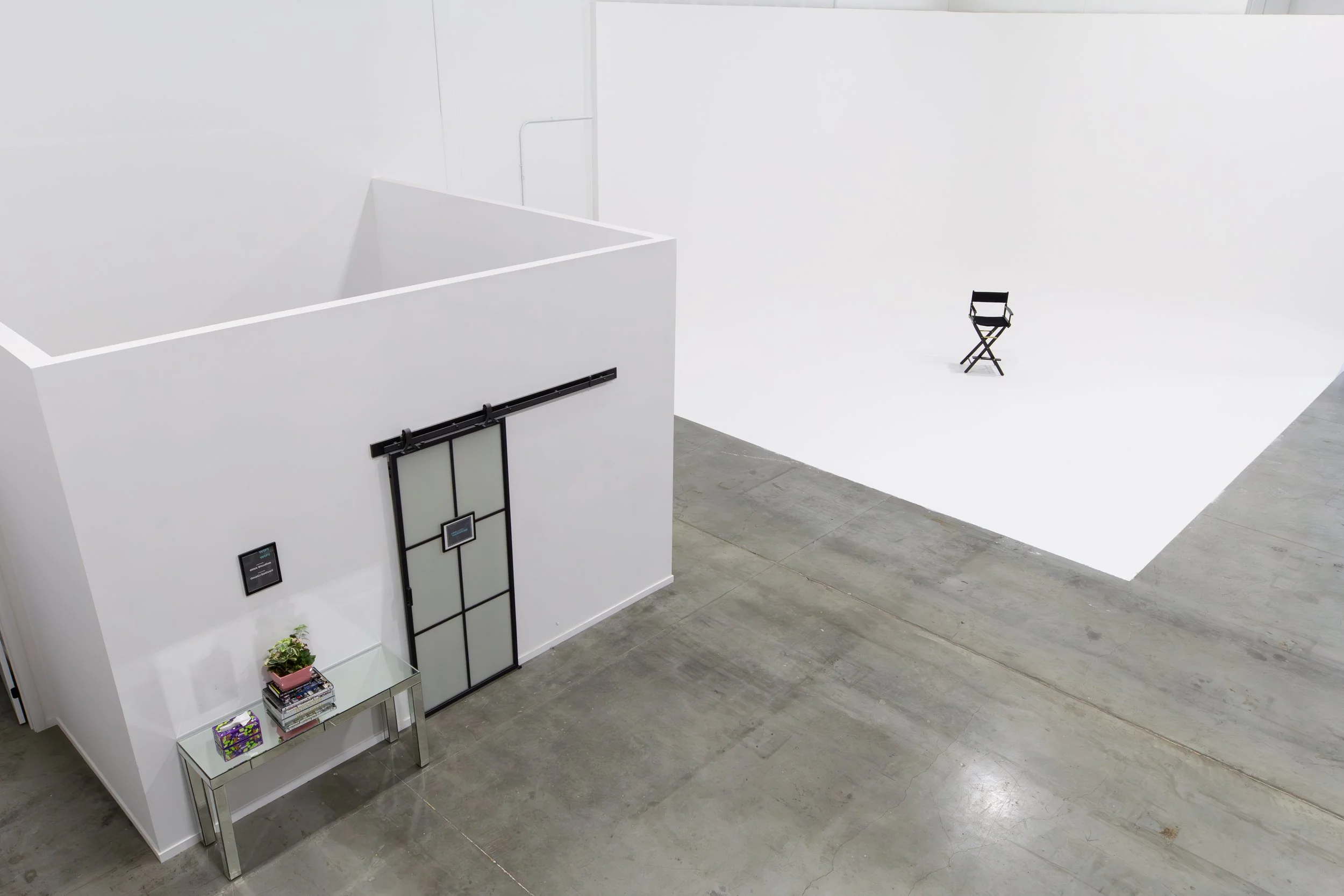 Art studio with a black director's chair on a white cyclorama backdrop, a sliding barn door, and a table with books, a potted plant, and a gift box. ONA Studios.