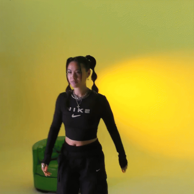 Young woman with long hair in pigtails smiling, wearing a black Nike crop top, black Nike pants, and a chain necklace, standing in front of a yellow wall with a green chair behind her.