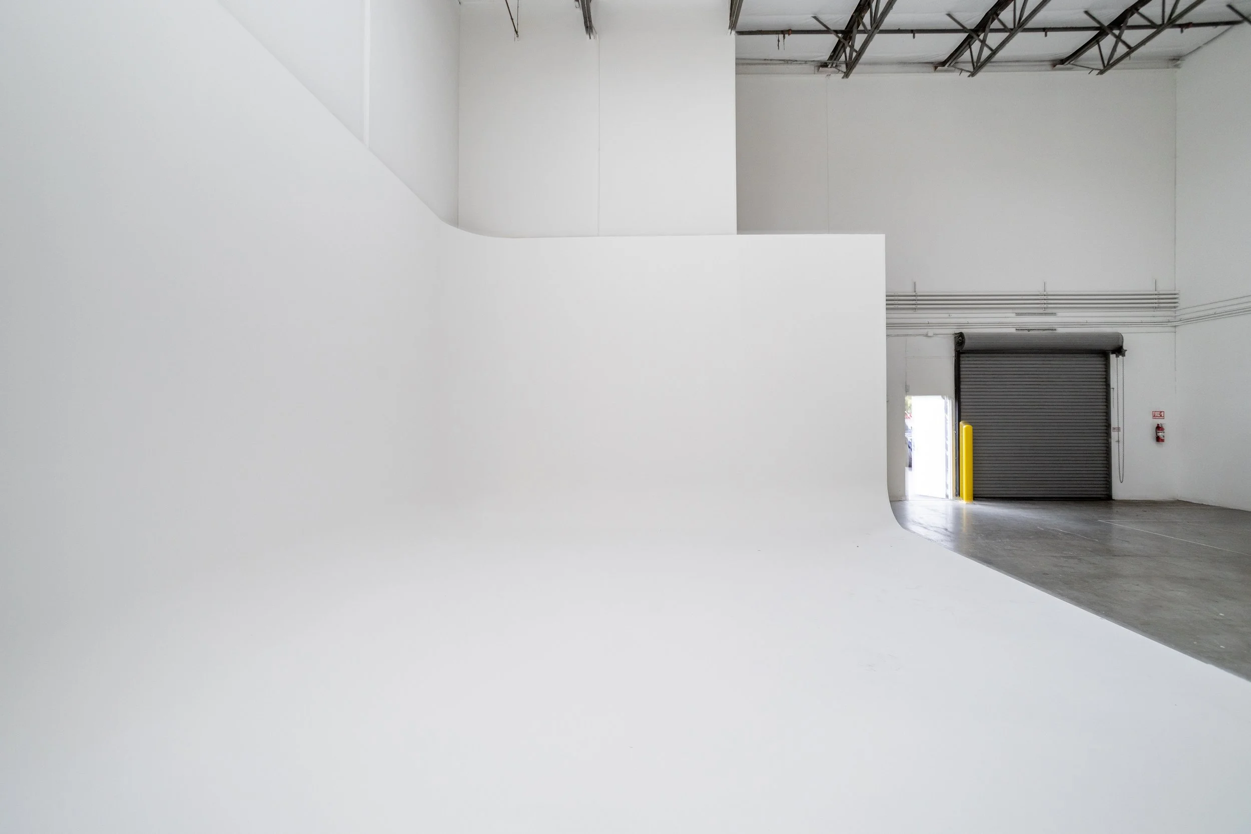 Interior of an empty warehouse or industrial space featuring white walls, high ceilings, a large closed roller door, and an open loading dock area. ONA Studios.