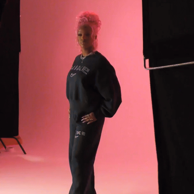 Monaleo with pink hair posing in a photo studio with a pink background, wearing a black Nike sweatshirt and matching black Nike pants.