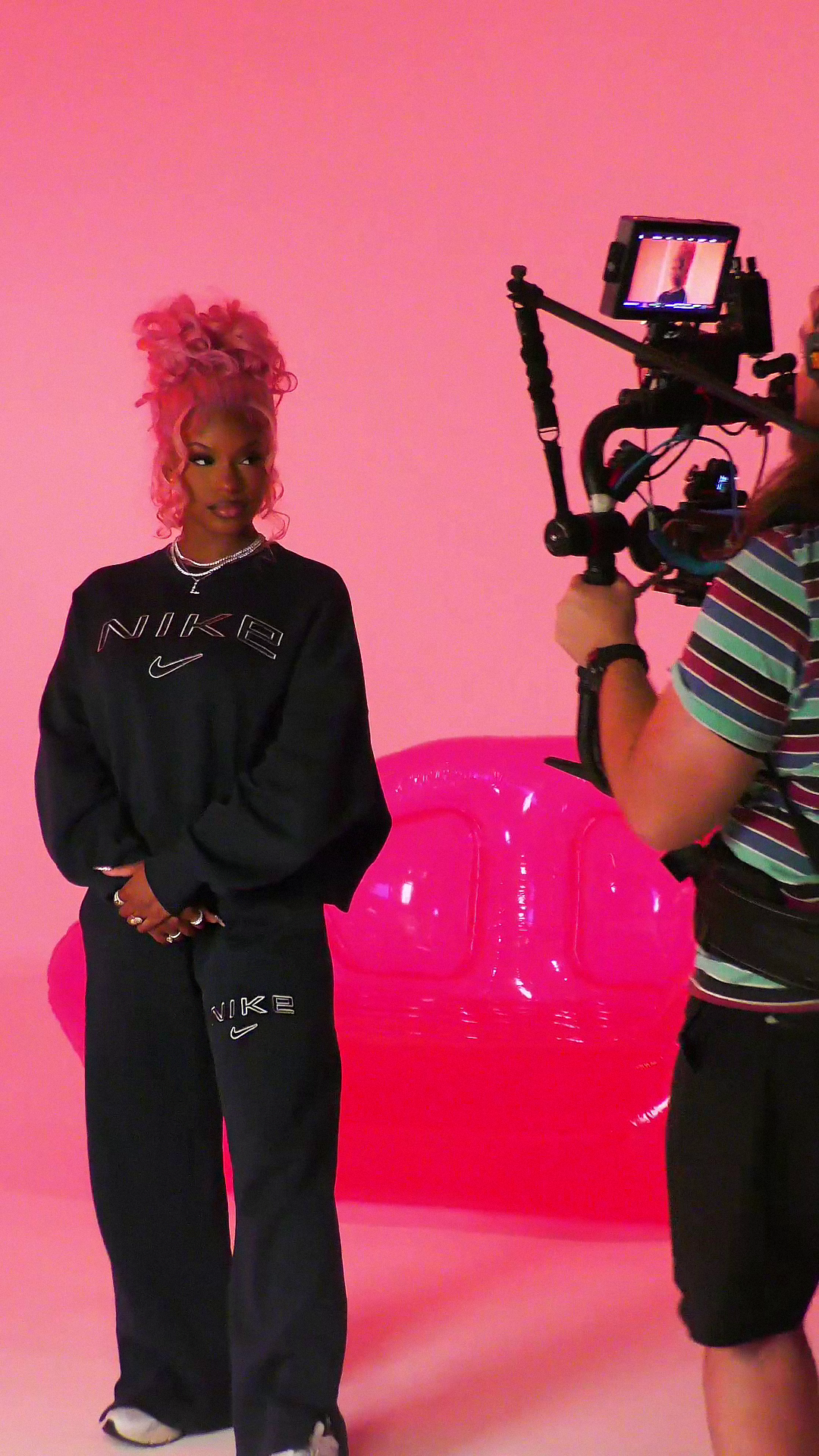 Monaleo wearing a black Nike sweatshirt and pants, standing in front of a pink background with a pink inflatable lips sculpture, while a person with a striped shirt is filming her with a video camera.