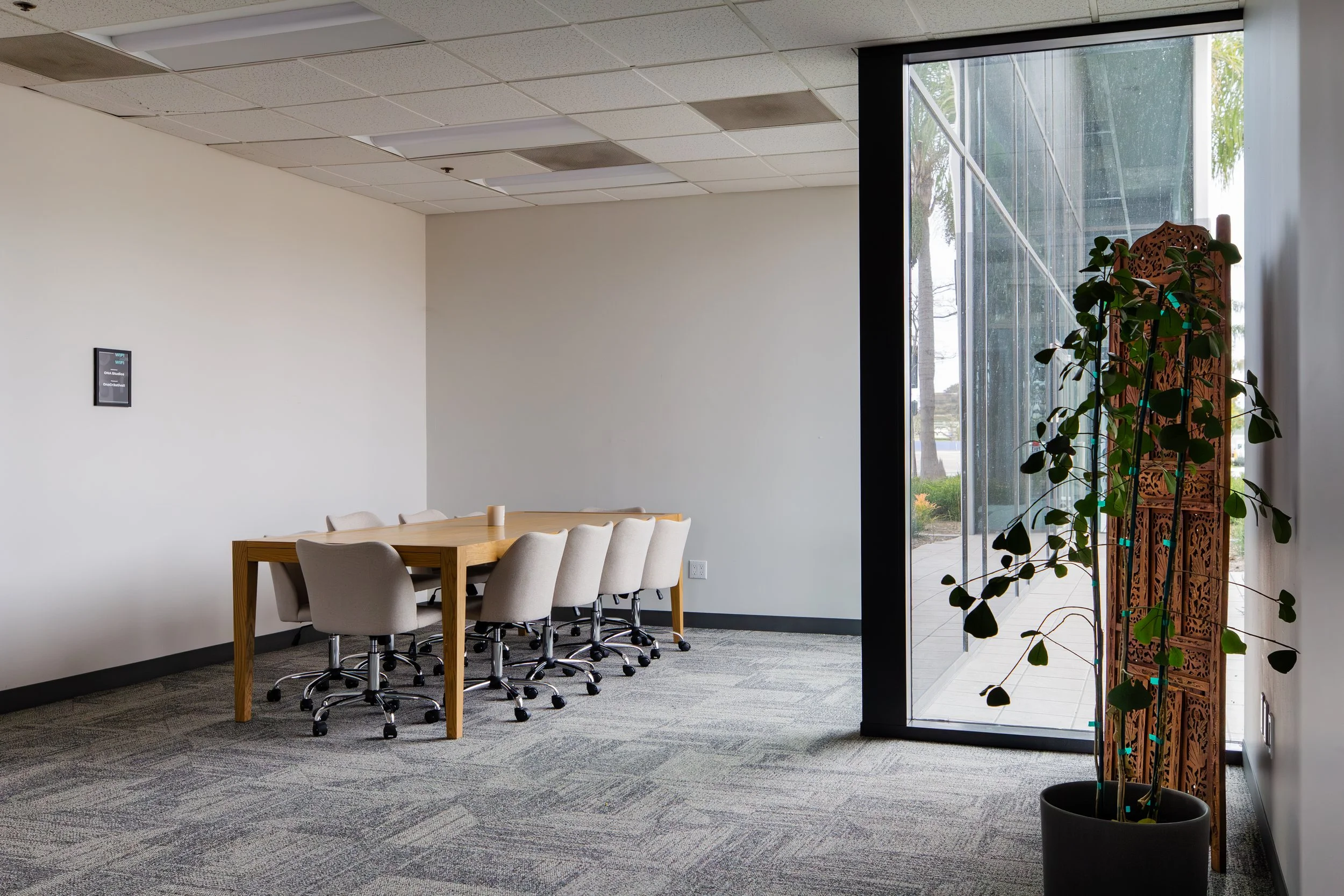 A conference room with a wooden table surrounded by beige office chairs on wheels. There is a large window with a glass wall showing trees outside, and a potted plant nearby. The ceiling has acoustic tiles, and the floor is carpeted. ONA Studios.