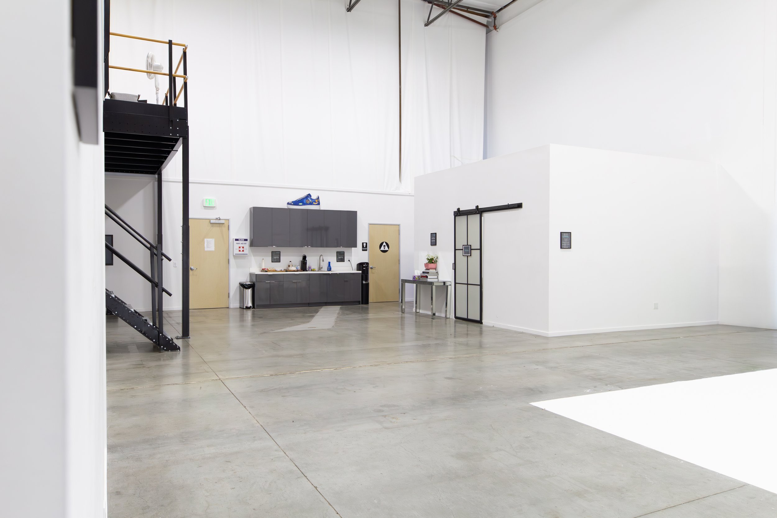 Inside a spacious, industrial-style room with white walls and a concrete floor, featuring a black staircase on the left, a kitchenette with gray cabinets on the back wall, and a small table with books and plants on the right. ONA Studios.