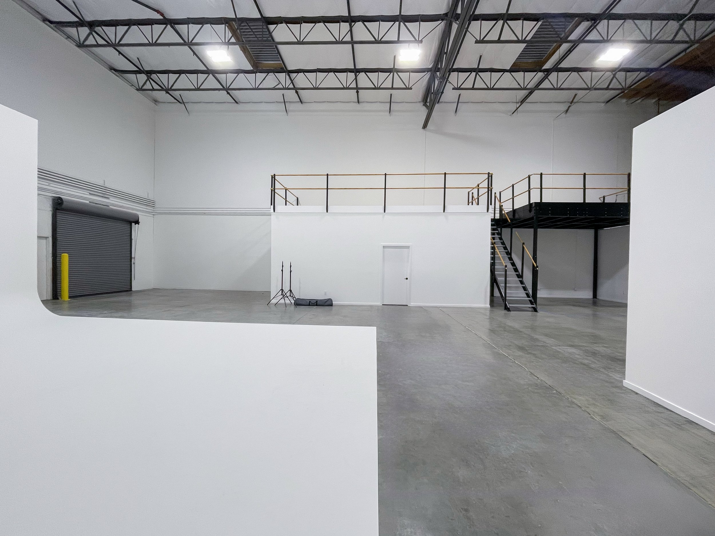 Empty industrial warehouse with white walls, concrete floors, a rolling metal door on the left, and a black metal staircase leading to a second-floor platform with safety railings. ONA Studios.