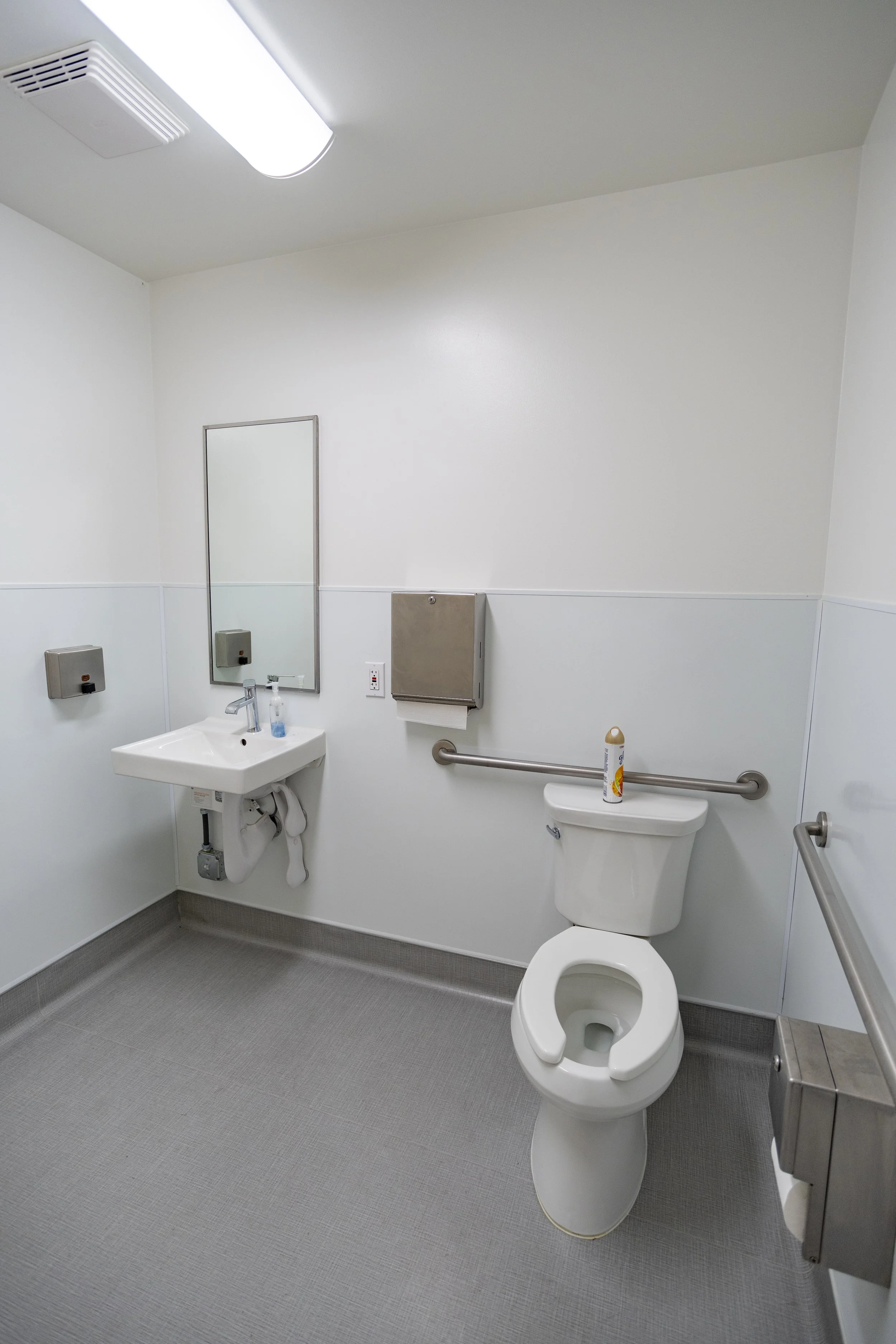 Empty bathroom with a toilet, small sink, mirror, hand sanitizer, soap dispensers, and grab bars. ONA Studios.