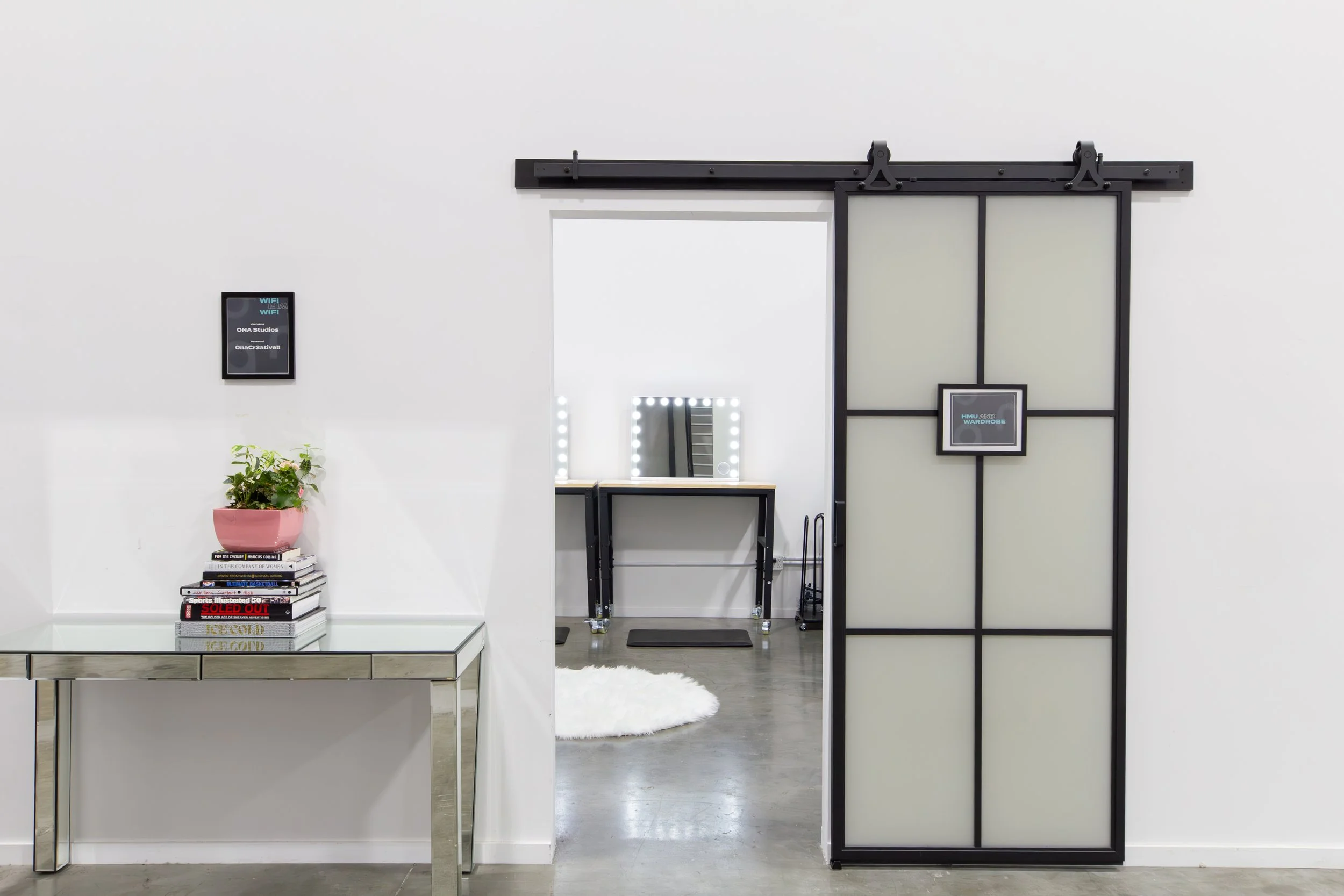 Modern interior with white walls, a metal and glass console table holding a potted plant and books, a black framed sliding door, a small digital display on the wall, and a dressing area with a mirror surrounded by lights, a table. ONA Studios.