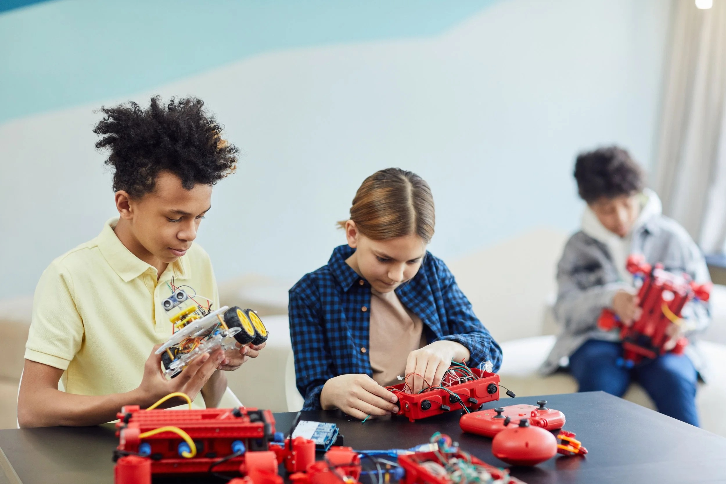 Three children working on robotics projects at a table, with electronic parts and controllers visible.