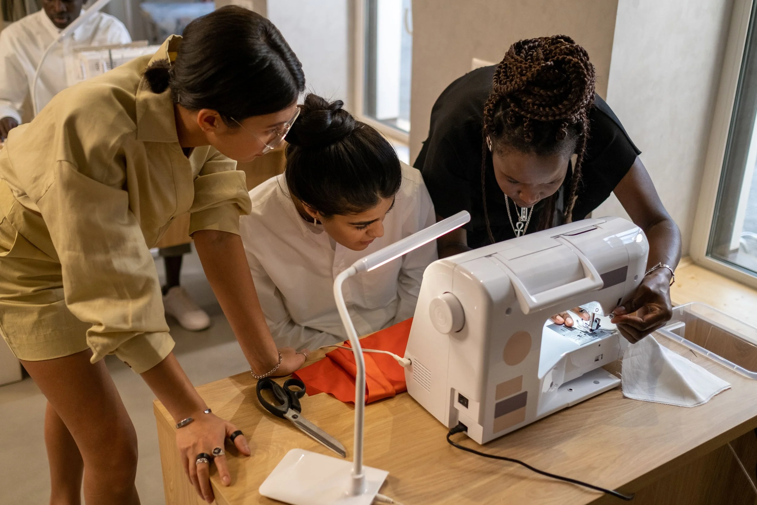 Three women are gathered around a sewing machine, closely examining and working on a fabric. Two women are standing, leaning over the sewing machine, while one woman is seated at the machine. The setting appears to be indoors, likely a sewing workshop or classroom.