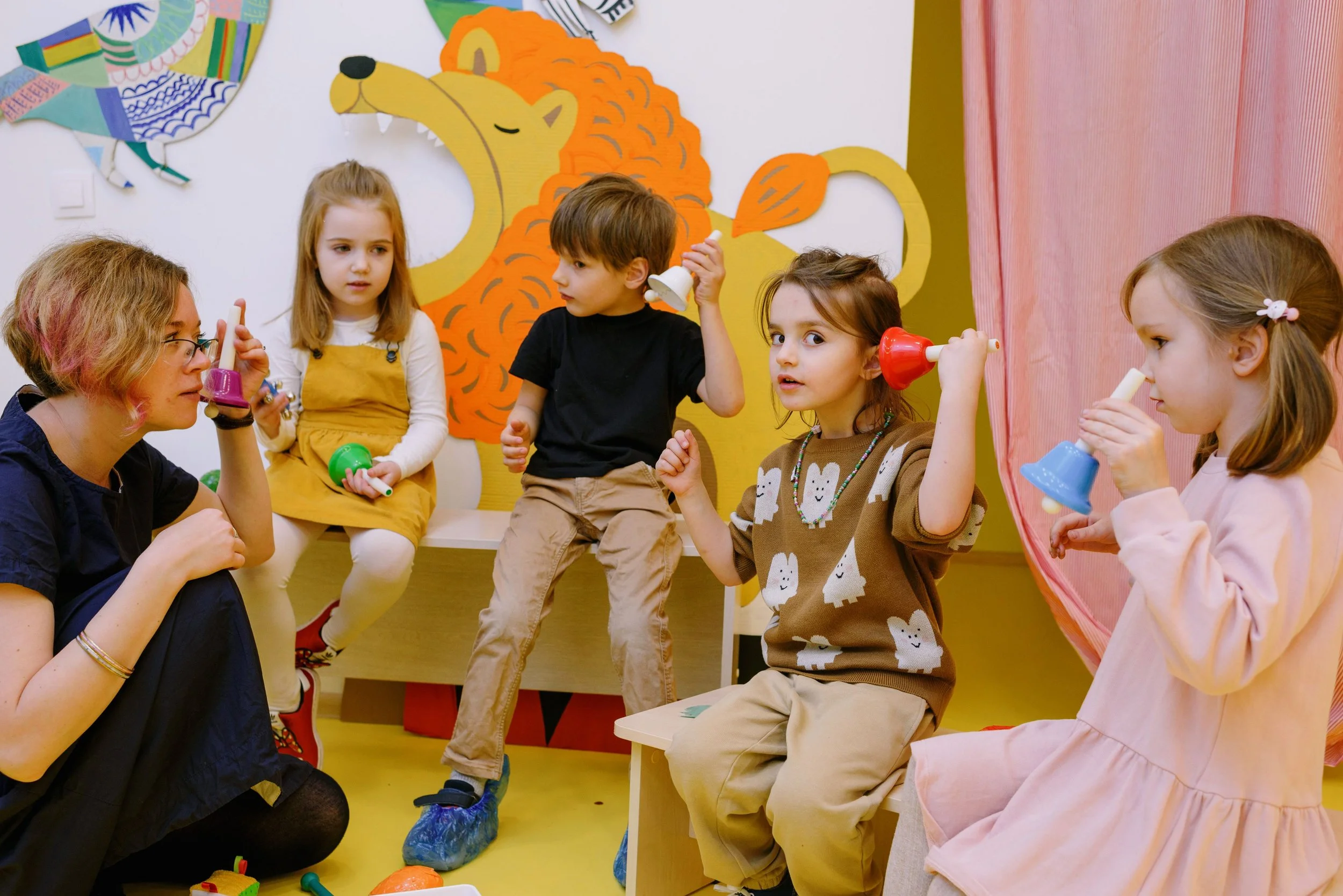 A group of young children and an adult woman in a colorful classroom. The children are holding small musical instruments and sitting on benches, with a large, smiling lion art piece on the wall behind them.