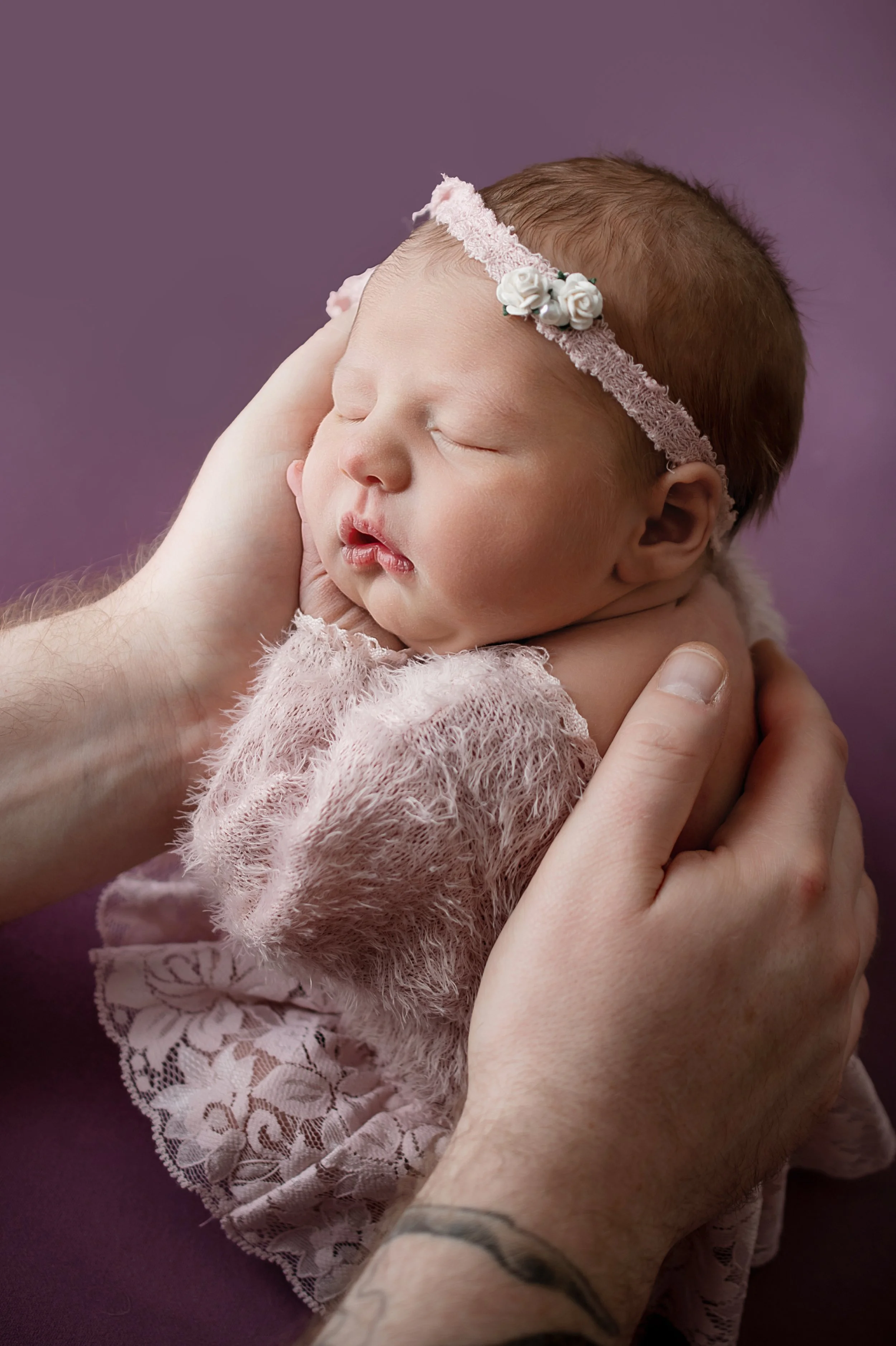 Close-up of a newborn baby girl sleeping peacefully, wearing a pink headband with white roses and a pink fuzzy dress, being gently held by an adult's hands against a purple background.