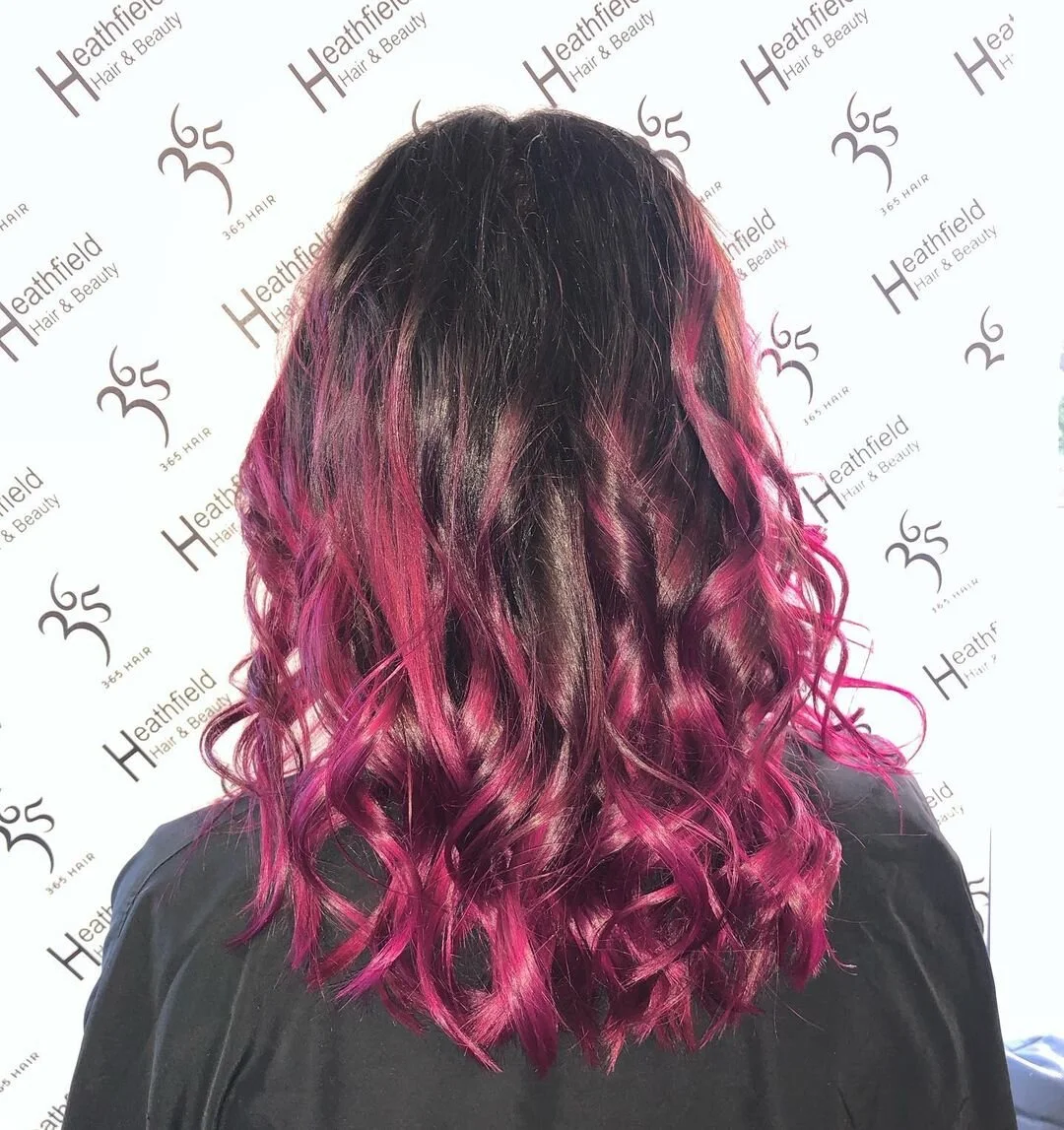 Back view of a woman with curly, shoulder-length black hair transitioning into bright pink ombre at the ends.