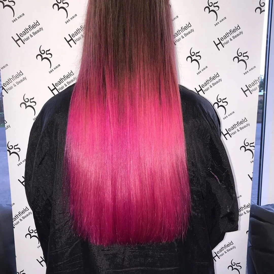 Back view of a person with long, straight hair that is dark at the top and transitions into vibrant pink towards the bottom, standing in front of a white background with black text and logos for a hair salon.