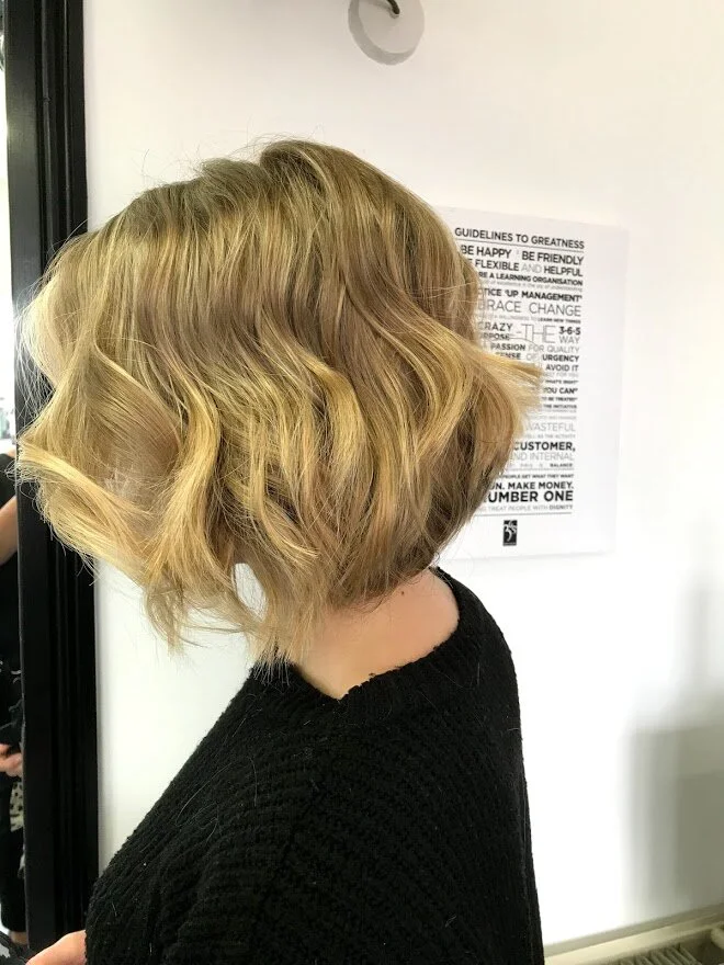 Side view of a person with a short, wavy blonde bob hairstyle wearing a black sweater, standing in front of a white wall with a printed sign.