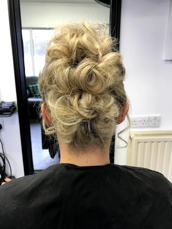 Back view of a woman with blonde, curly hair styled in an elegant updo, sitting in a salon.