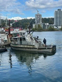 A small tugboat docked on a river with a person standing on the boat. In the background are city buildings, cranes, and trees under a partly cloudy sky. KNW Diesel provide installation, sales and service of Marine engines Scania, FPT, Moteurs Baudoui