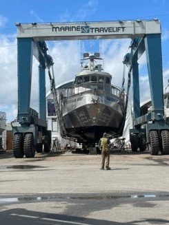 Large boat being lifted out of the water by a Marine Travelift crane at a shipyard, with a worker standing nearby KNW Diesel provide installation, sales and service of Marine engines Scania, FPT, Moteurs Baudouin in British Columbia