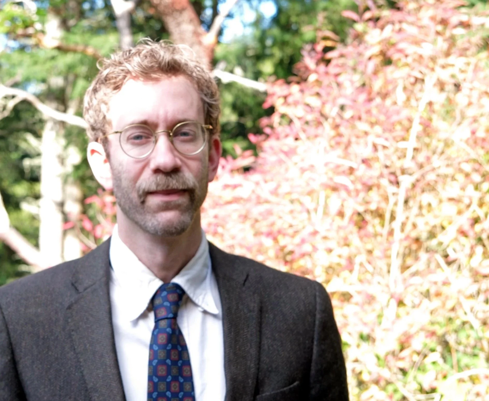 A man with curly hair, glasses, and a beard wearing a suit and tie, standing outdoors with trees and pink flowering bushes in the background.