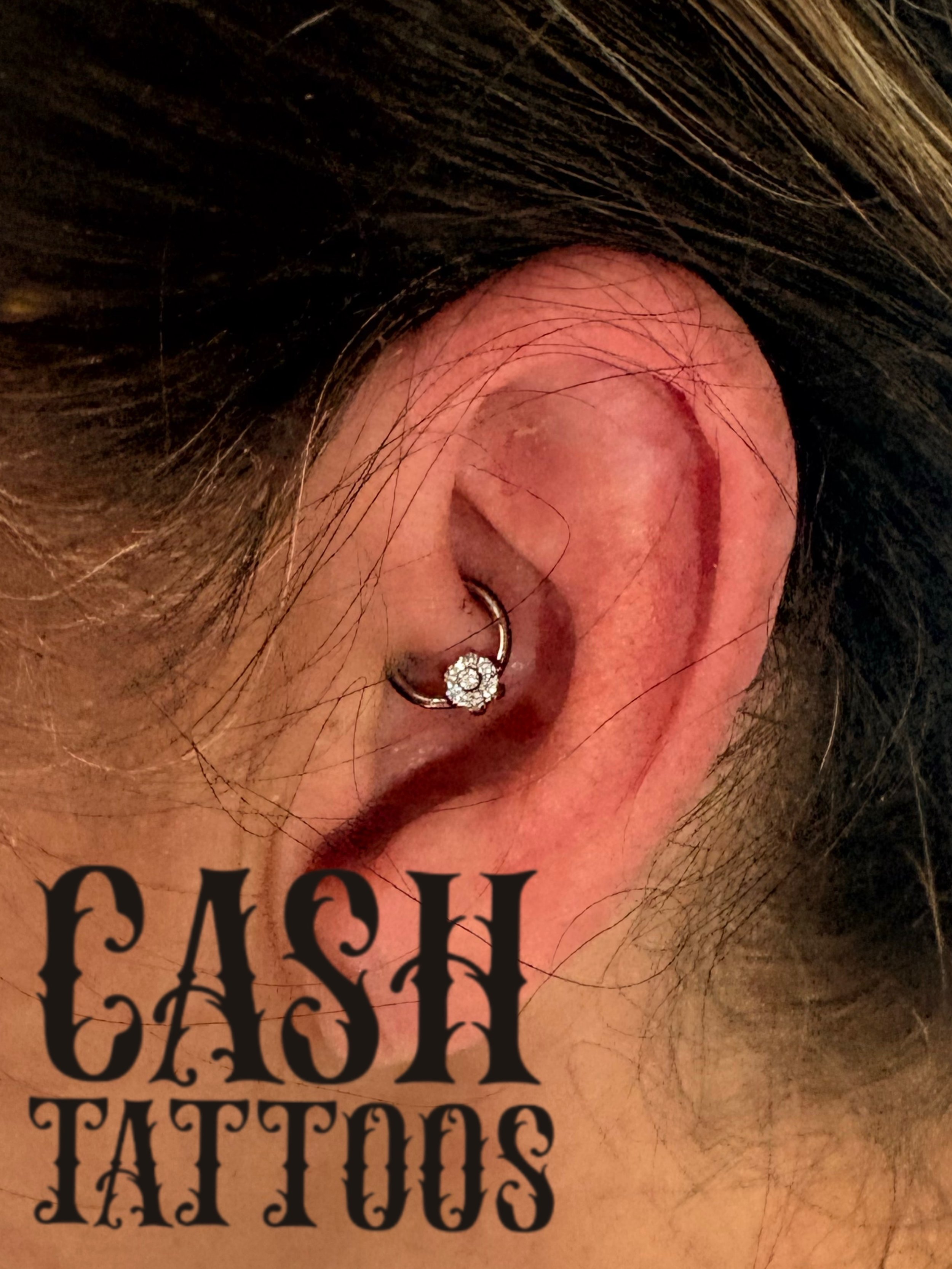 Close-up of a person's ear with a hoop earring and a small tattoo that reads 'CASH TATTOOS' in bold black letters.