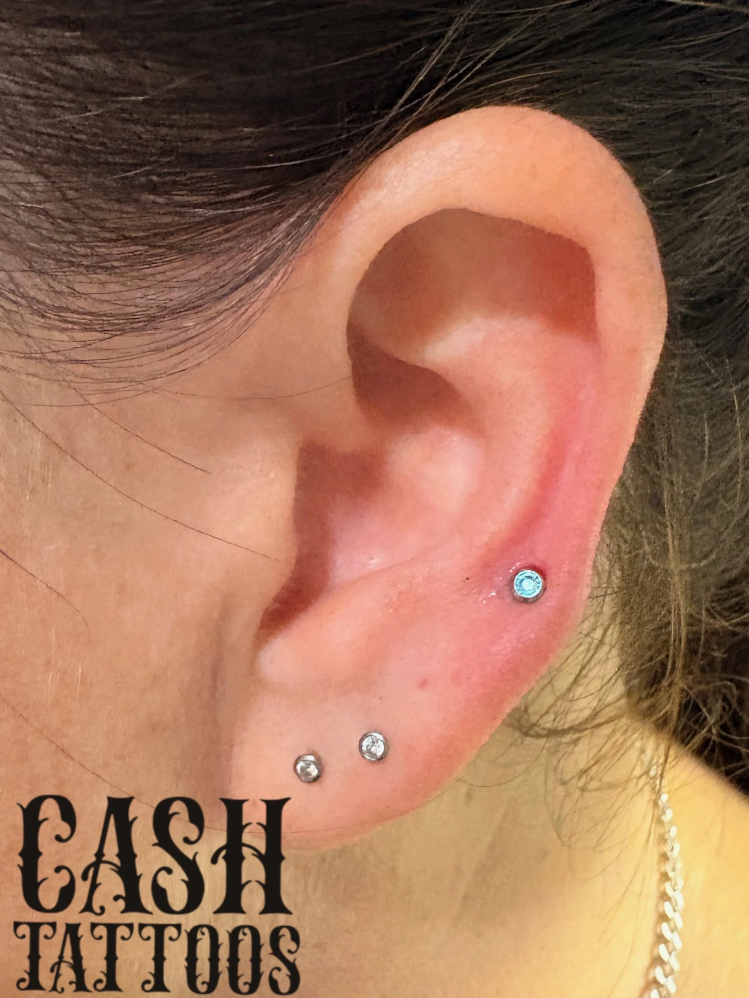 Close-up of a person's ear with three piercings, one with a small blue gemstone in the upper cartilage and two with small clear gemstones on the lobe, with a text overlay reading 'CASH TATTOOS'.