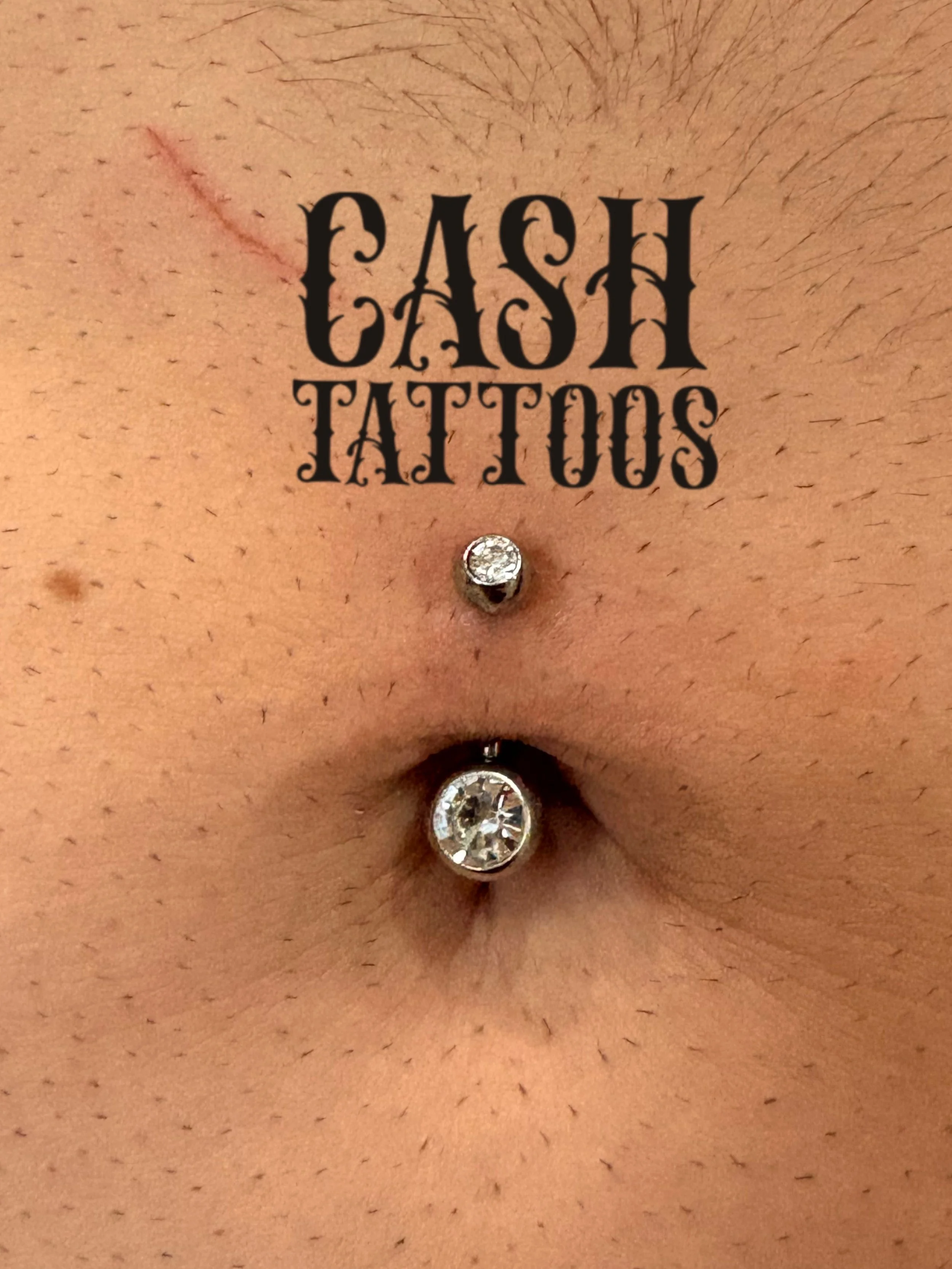 Close-up of a person's navel with a tattoo that says 'CASH TATTOOS' and two embedded diamond-like jewelry piercings.