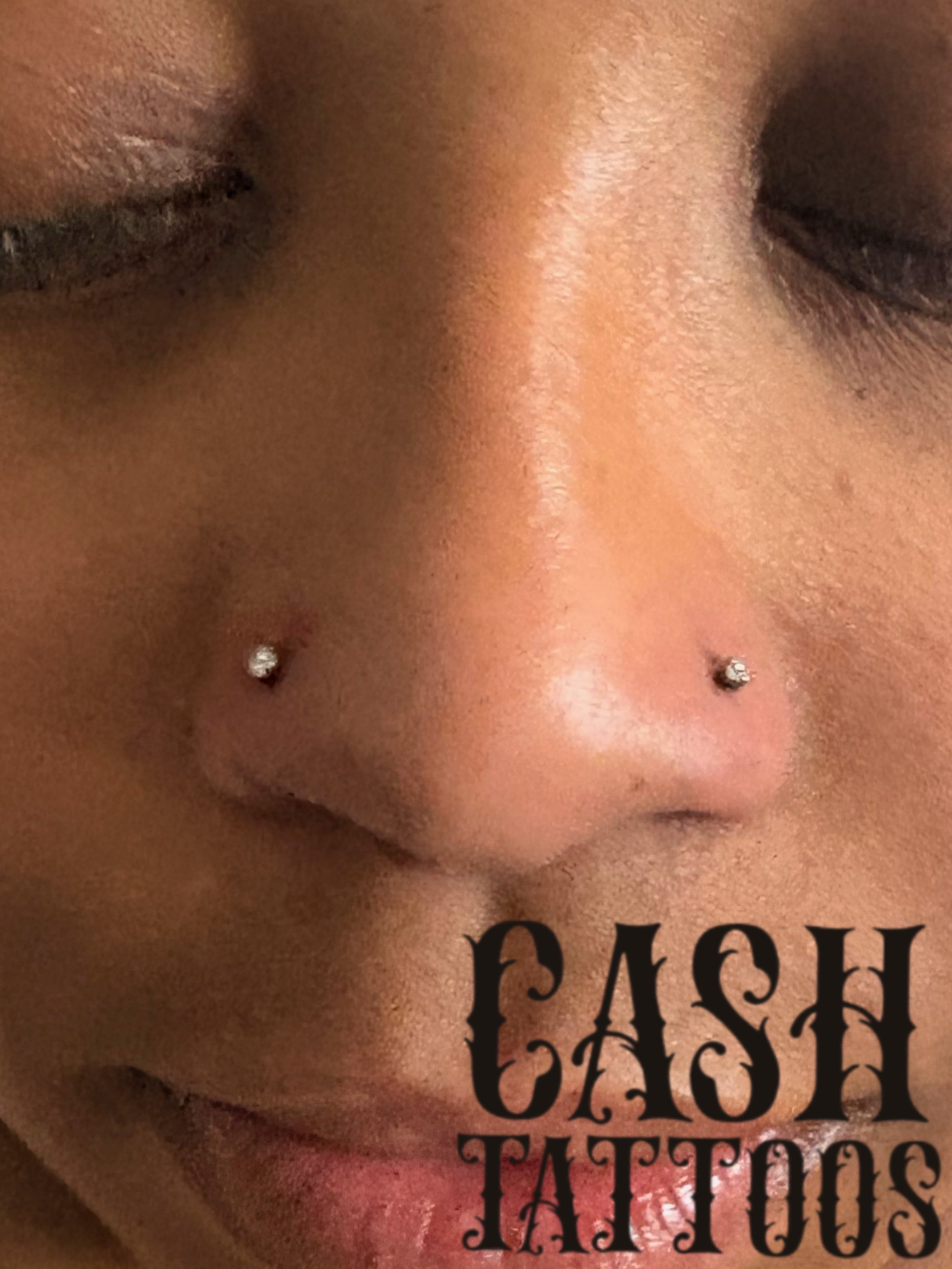 Close-up of a person's face with closed eyes, featuring two small nose piercings and the words 'CASH TATTOOS' in stylized black font over the chin.