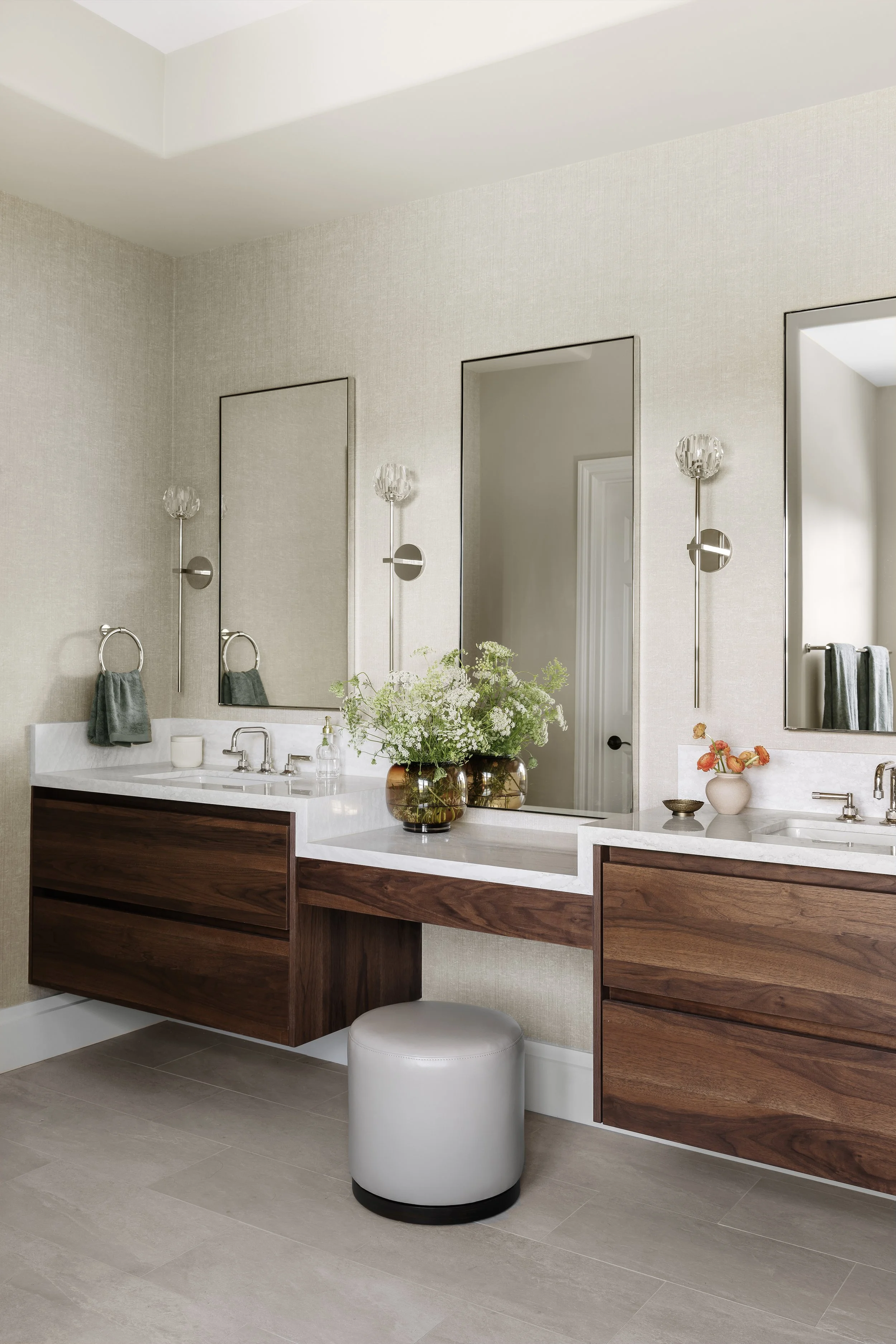 Modern bathroom with double vanity, large mirrors, wall-mounted light fixtures, a vase with flowers, and a small cushioned stool.