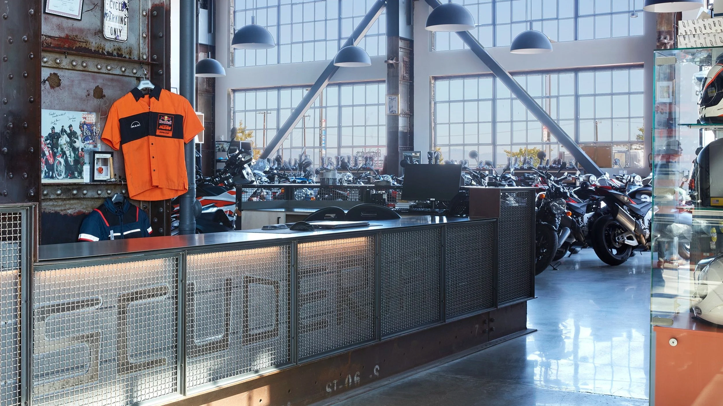 Inside a motorcycle showroom with a reception desk, display of racing jerseys on the wall, and multiple motorcycles parked near large glass windows letting in natural light.