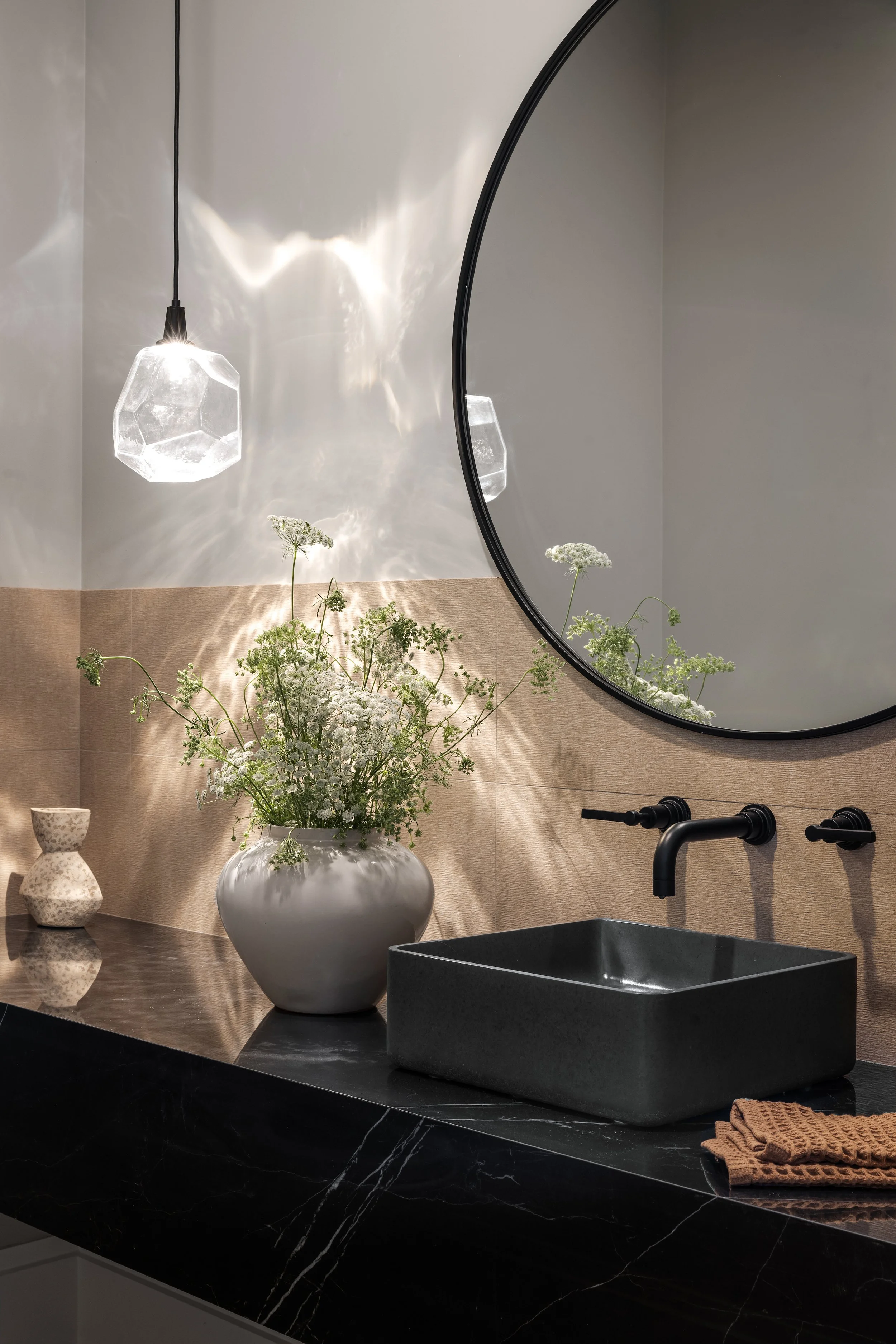 A modern bathroom with a black square vessel sink, black faucet, round mirror, and a hanging geometric glass pendant light. A white vase with green plants and white flowers, a small textured ceramic vase, and a folded brown towel sit on black marble 